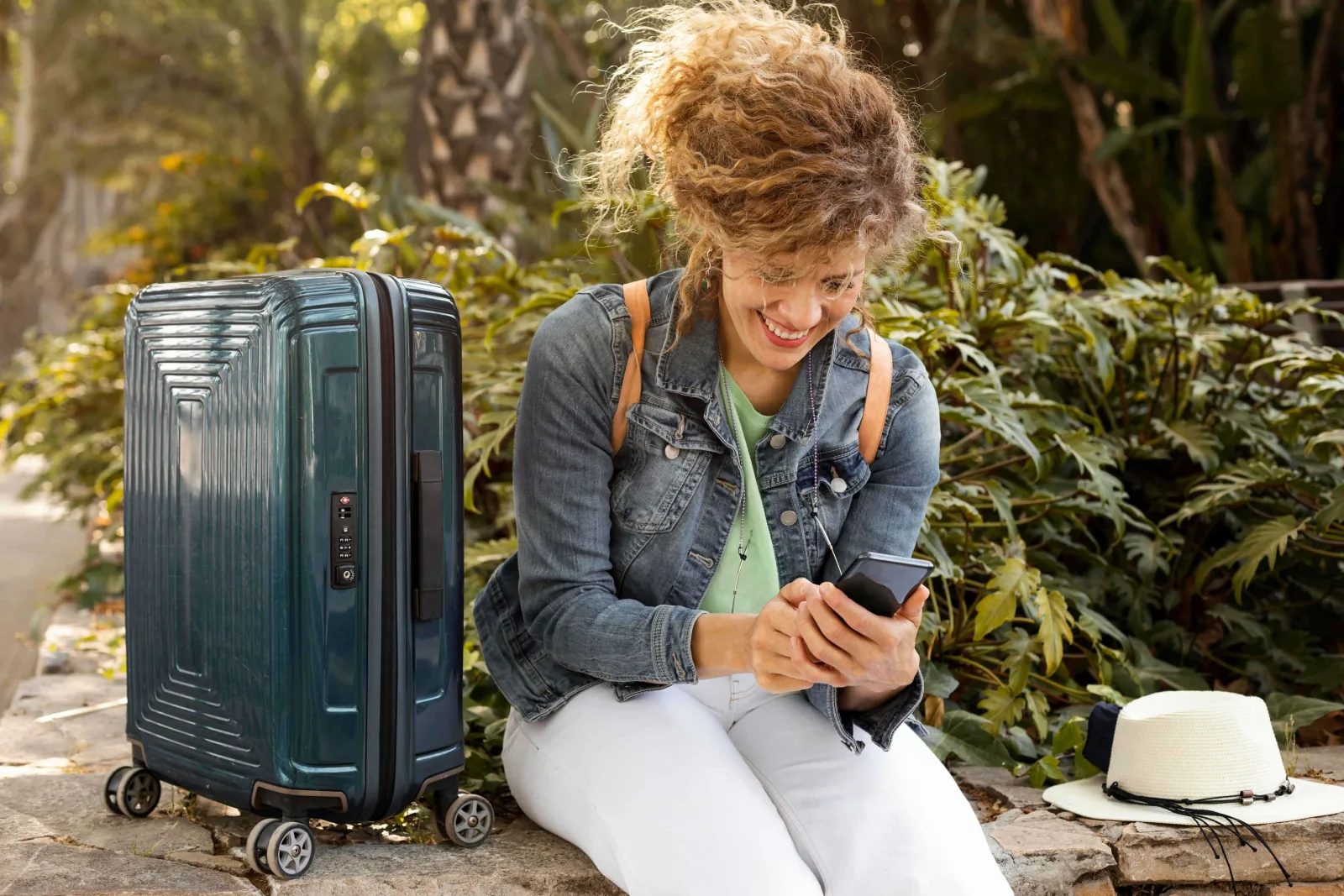 Smiling young woman setting up her swap project on her phone, suitcase beside her