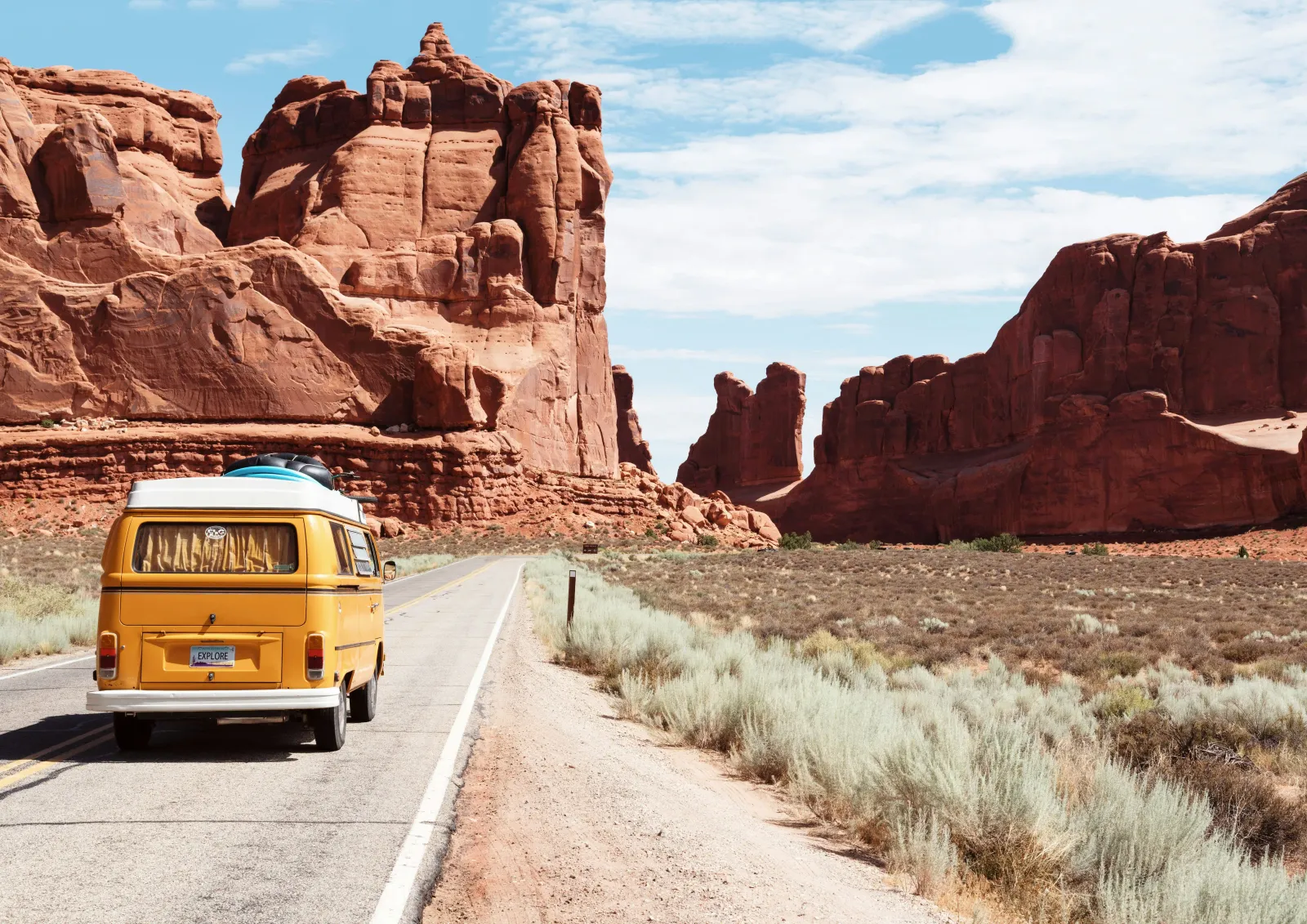 Combi Volkswagen jaune sur une route déserte face à des paysages de roches rouges, plaque « EXPLORE »